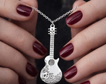 May include: A silver guitar pendant necklace. The guitar pendant has a detailed design, including a skull and floral pattern. The necklace is held by hands with dark red nail polish, against a dark background.