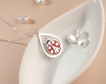 May include: A silver necklace with a teardrop pendant featuring a red and black ladybug design. The necklace is displayed on a white, leaf-shaped dish, accompanied by several white pearls. The background is a soft, neutral color.