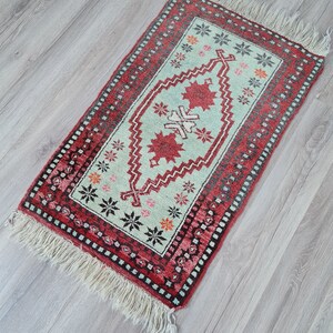 May include: A small, rectangular rug with a traditional Turkish design. The rug has a light green background with a red border and intricate patterns in black, white, and orange. The rug has fringe on all four sides.