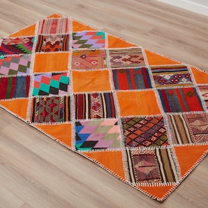 Puede incluir: Una alfombra patchwork naranja con forma de diamante. La alfombra presenta una variedad de cuadrados coloridos y estampados cosidos entre sí. Los bordes están acabados con costuras blancas, creando un revestimiento de suelo vibrante y texturizado.