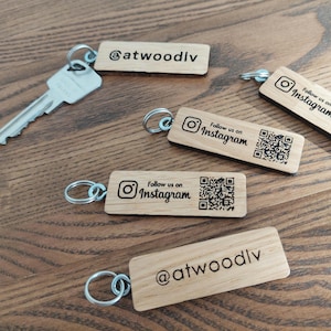 Könnte beinhalten: Holz-Schlüsselanhänger mit silbernen Schlüsselringen. Einige Schlüsselanhänger tragen den Text "Follow us on Instagram" mit einem QR-Code. Andere tragen den Text "@atwoodlv". Die Schlüsselanhänger liegen auf einer Holzoberfläche.