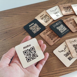 May include: Square wooden QR code signs with various designs. Some signs are black, others are light wood. Text includes "menu", "Table No. 7", "Table No. 8", and social media handles. The signs are for a restaurant or bar.