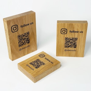 Peut inclure: Trois blocs de bois avec le logo Instagram, les mots "follow us", un code QR et le nom d'utilisateur "@atwoodlv" imprimés en noir. Les blocs sont en bois brun clair et sont disposés sur une surface blanche.