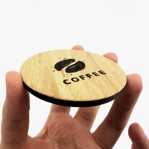May include: A round wooden coaster with a black rim, featuring a coffee bean design and the word "COFFEE" in a simple font. The coaster is held in a hand against a white background.