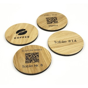 Op de afbeelding: Vier houten onderzetters met donkere randen. Eén onderzetter heeft een koffieboonafbeelding en het woord "COFFEE". Een andere zegt "Coffee Shop" met een QR-code en "Menu". De derde zegt "Table #14". De laatste zegt "Scan for Menu" met een QR-code en "Table Nr. 8".