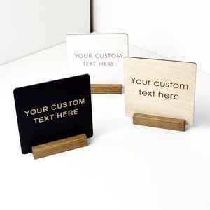 May include: Three square signs with wooden bases. One sign is black, one is white, and one is natural wood. Each sign has the text "YOUR CUSTOM TEXT HERE" engraved on it. The signs are displayed on a white surface.