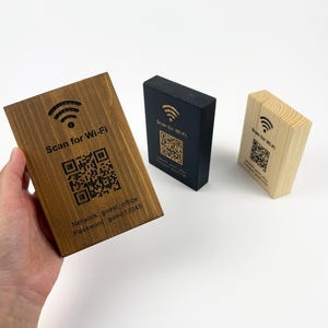 Può includere: Tre cartelli in legno per il Wi-Fi. Il più grande è tenuto in mano. Ogni cartello ha un codice QR e la scritta "Scan for Wi-Fi". Uno è in legno scuro, uno nero e uno in legno chiaro.