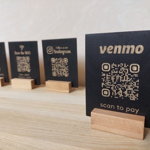 May include: Five black square signs with QR codes and text on them. The signs are on wooden stands. The text on the signs reads "Black Bison", "Scan for Wifi", "Follow us on Instagram", "venmo", and "scan to pay".