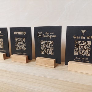 May include: Four black square signs with QR codes and text on them. The signs are on wooden stands. The text on the signs reads: "Black Bison", "venmo", "Follow us on Instagram", and "Scan for WiFi". The sign with "Scan for WiFi" also has the text "Name: GUEST Password: 12345".