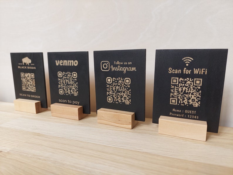 May include: Four black square signs with QR codes and text on them. The signs are on wooden stands. The text on the signs reads: "Black Bison", "venmo", "Follow us on Instagram", and "Scan for WiFi". The sign with "Scan for WiFi" also has the text "Name: GUEST Password: 12345".