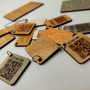 May include: A collection of wooden keychains with laser-engraved QR codes and the text "SCAN ME". The keychains are made of different shades of wood and have a natural finish.