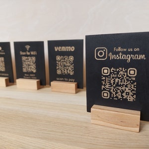 May include: Four black wooden signs with QR codes and text. The signs are on a wooden table. The text on the signs reads "Black Bison", "Scan for WiFi", "venmo scan to pay", and "Follow us on Instagram".