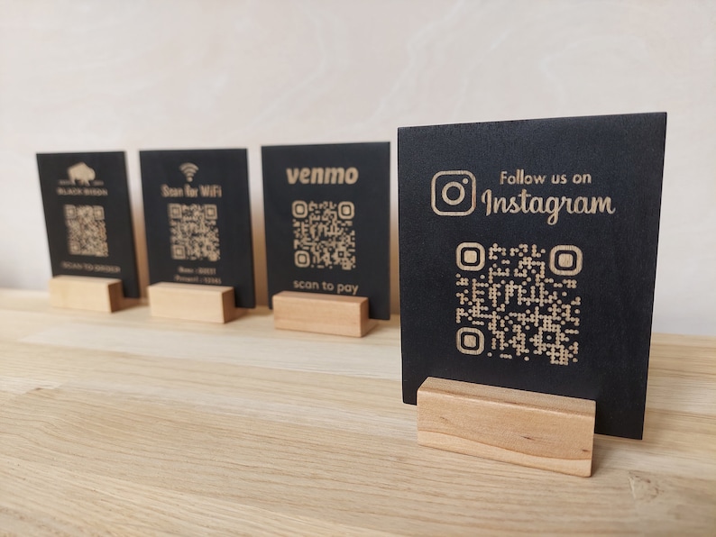 May include: Four black wooden signs with QR codes and text. The signs are on a wooden table. The text on the signs reads "Black Bison", "Scan for WiFi", "venmo scan to pay", and "Follow us on Instagram".