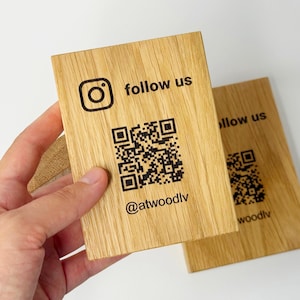 May include: Wooden rectangular sign with the Instagram logo, the words "follow us", a QR code, and the username "@atwoodlv" printed in black. The sign is held in a hand and another sign is in the background.