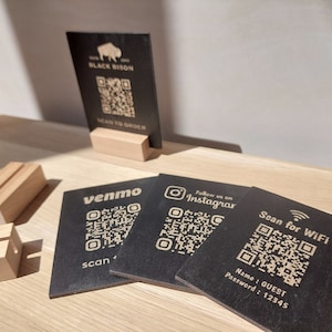 May include: Four black square cards with QR codes and text. The cards are on a wooden surface. The text on the cards reads "Black Bison", "venmo", "Follow us on Instagram", and "Scan for WiFi". The card with "Scan for WiFi" also includes the text "Name: GUEST Password: 12345".