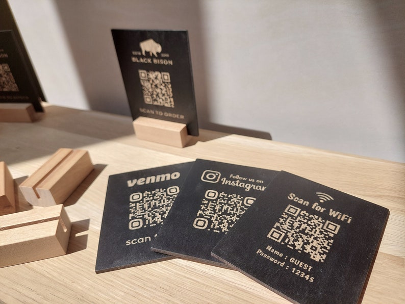 May include: Four black square cards with QR codes and text. The cards are on a wooden surface. The text on the cards reads "Black Bison", "venmo", "Follow us on Instagram", and "Scan for WiFi". The card with "Scan for WiFi" also includes the text "Name: GUEST Password: 12345".