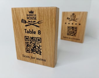 Op de afbeelding: Een houten tafelnummerbord met een zwart-witte QR-code, de tekst "Table 8" en de woorden "Scan for menu". Het bord heeft ook een logo met de tekst "Barbecue House" en een kroon, een grill, een vork en een spatel.