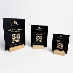 Può includere: Tre cartelli rettangolari neri con basi in legno. Ogni cartello presenta il testo "The Green Fork" e "Leave us a review on Google" con un codice QR. I cartelli sono di diverse dimensioni.