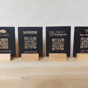 May include: Four black square signs with gold text and QR codes. The signs are on wooden stands. The signs read "Black Bison", "Venmo", "Follow us on Instagram", and "Scan for WiFi".