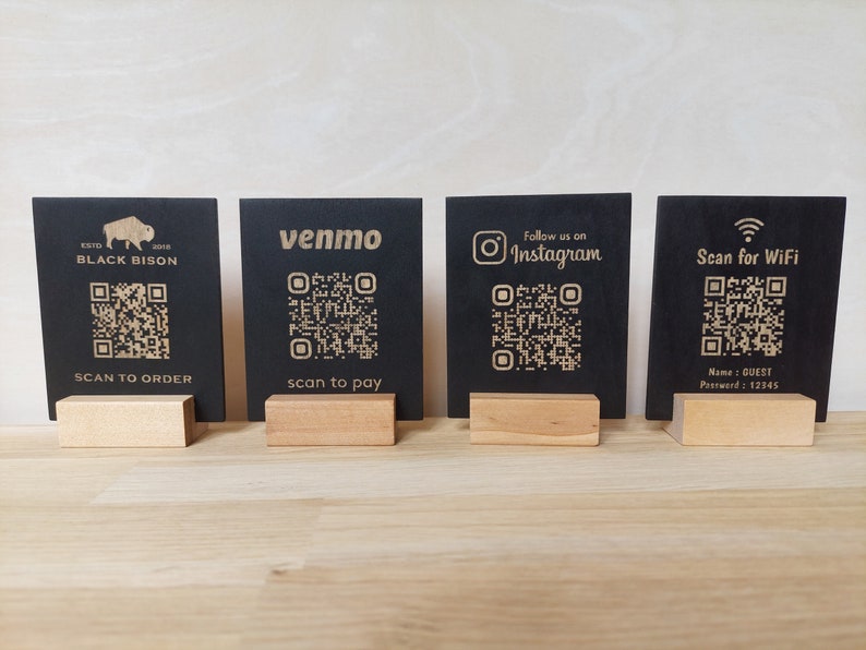 May include: Four black square signs with gold text and QR codes. The signs are on wooden stands. The signs read "Black Bison", "Venmo", "Follow us on Instagram", and "Scan for WiFi".