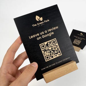 Puede incluir: Cartel de madera negro con el texto "The Green Fork Eat Well" y "Leave us a review on Google" en color claro. Un código QR aparece debajo del texto. El cartel está sujeto por una base de madera.
