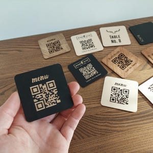 Op de afbeelding: Een set houten onderzetters met verschillende ontwerpen, waaronder een QR-code, de woorden "Table No. 8" en het woord "Menu". De onderzetters zijn gemaakt van lichtbruin hout en hebben een zwart-wit ontwerp.