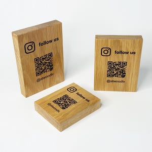 Può includere: Tre blocchi di legno rettangolari con il logo di Instagram, le parole "follow us", un codice QR e il nome utente @atwoodlv stampati in nero. I blocchi sono in legno marrone chiaro.