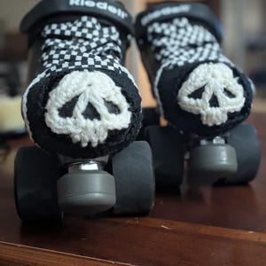 May include: Pair of black roller skates with white and black checkered laces. Each skate has a crocheted skull design in white on the front. The skates are on a dark brown wooden surface.