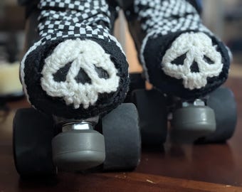 May include: Pair of black roller skates with white and black checkered laces. Each skate has a crocheted skull design in white on the front. The skates are on a dark brown wooden surface.