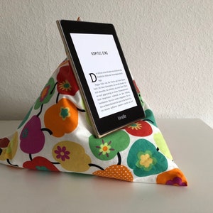 May include: A white pillow with a colourful floral pattern is shaped like a pyramid and holds a Kindle e-reader. The Kindle screen displays the text "KAPITEL EINS" and "Kindle".
