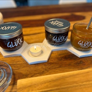 Könnte beinhalten: Drei gläserne Gläser mit Deckeln, jedes mit einer anderen Art von Marmelade gefüllt. Die Gläser sind mit "Glück" beschriftet und haben unterschiedliche Geschmacksbeschreibungen. Die Gläser sind auf einem weißen sechseckigen Tablett mit einer kleinen weißen Kerze in der Mitte angeordnet.