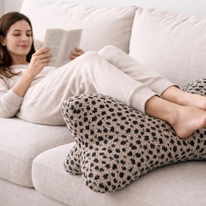 May include: A patterned reading pillow in beige and black sits on a light-coloured sofa. The pillow has a distinctive shape, designed for comfort. The image shows a person relaxing on the sofa with the pillow.