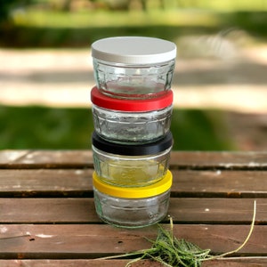 May include: Stack of four glass jars with lids. The top jar has a white lid, the second jar has a red lid, the third jar has a black lid, and the bottom jar has a yellow lid.
