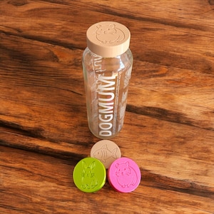 May include: A clear glass bottle with a beige lid featuring a hamster design. The bottle has the word "DOGMUM" printed vertically. Three round, colourful tokens with animal designs are also present: a fish, a rabbit, and a hamster.