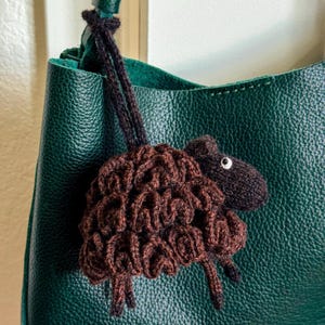 May include: A dark brown crocheted sheep ornament with a textured, curly wool body and a smooth head featuring a single white eye. The sheep is attached to a dark green leather handbag by a thin, dark brown crocheted cord loop.
