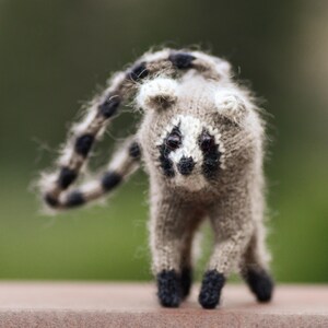 May include: A small, hand-knitted lemur figurine crafted from fuzzy, light brown yarn. It features a distinct white face mask with black patches around the eyes, a small black nose, and a long, curved tail with alternating black and brown rings.