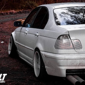 May include: A white saloon car with black tinted windows and aftermarket wheels. The car is parked on a dirt road with trees in the background. The car has a number plate that reads "LT".