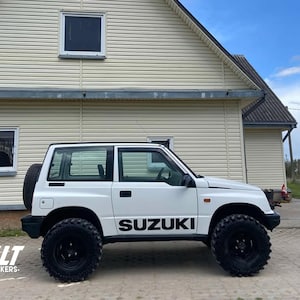 Puede incluir: Un SUV Suzuki blanco con ruedas y neumáticos negros está estacionado frente a una casa. La palabra "SUZUKI" se muestra en letras negras en el lateral del vehículo. El vehículo tiene una rueda de repuesto montada en la parte trasera.