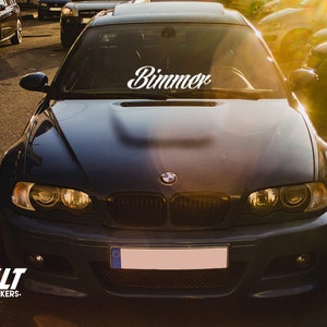Op de afbeelding: Een zwarte BMW met een witte "Bimmer" sticker op de voorruit. De auto staat geparkeerd op een parkeerplaats met andere auto's op de achtergrond.