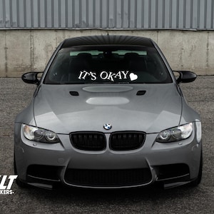 May include: A gray BMW car with a black grill and a white vinyl decal on the windshield that says "It's Okay" with a heart.