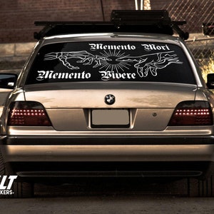 May include: A silver car with a black roof rack and a white decal on the rear window that reads "Memento Mori Memento Vivere" with a skeletal hand design.