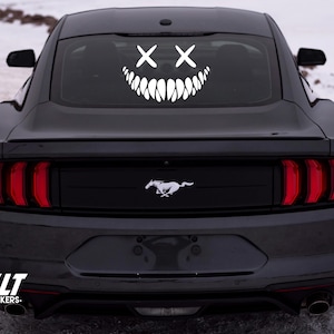 Può includere: Un'auto nera con un adesivo bianco di un viso sorridente con gli occhi incrociati sul lunotto posteriore. L'adesivo ha un ampio sorriso con i denti. L'auto ha un logo Ford Mustang sul retro.