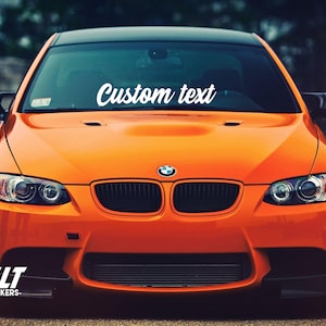 Può includere: Auto BMW arancione con un adesivo di testo personalizzato bianco sul parabrezza. Il testo recita "Custom text".
