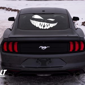 Può includere: Una Ford Mustang nera con un adesivo in vinile bianco di un viso sorridente sul lunotto posteriore. L'adesivo ha denti affilati e un'espressione minacciosa.