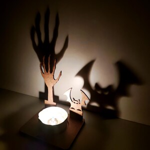 May include: A Halloween-themed candle holder with a wooden bat and hand silhouette. The candle holder has a square base with a lit candle, casting shadows of a hand and bat on the wall. The wooden cutouts are brown.