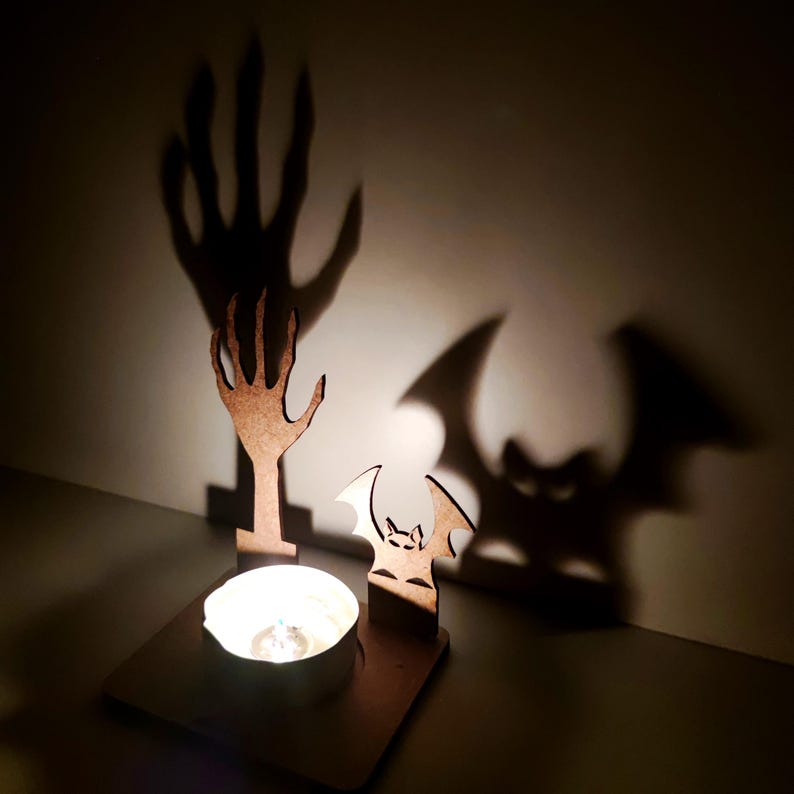 May include: A Halloween-themed candle holder with a wooden bat and hand silhouette. The candle holder has a square base with a lit candle, casting shadows of a hand and bat on the wall. The wooden cutouts are brown.