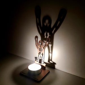 May include: A wooden candle holder featuring a cutout figure with arms raised, casting a shadow on the wall. The design includes a square base and a circular space for a tea light candle. The shadow mimics the figure's pose.