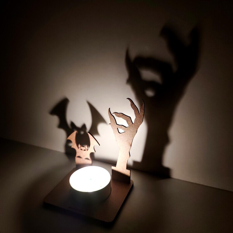 May include: A wooden Halloween-themed candle holder with a bat and a hand silhouette. The candle holder has a square base with a circular space for a tea light. The shadows cast by the figures create a spooky effect.