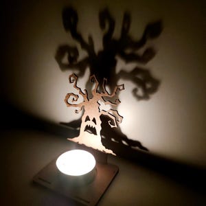 May include: A Halloween-themed candle holder featuring a spooky tree design. The wooden cutout casts a ghoulish shadow on the wall. A lit tea light sits in front of the tree, creating a warm glow. The tree has a carved face.