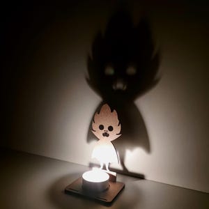 May include: A wooden candle holder with a cartoon ghost design. The candle holder casts a shadow of a larger ghost figure on the wall. The candle is lit, creating a warm glow and enhancing the shadow effect.